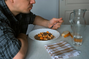 Man is eating buckwheat and chicken sitting at table at home, side view. Healthy food of poor person in third world country. Decanter and glass of water and two slices of bread on table.