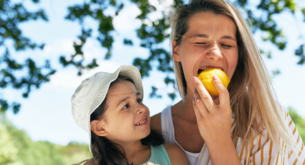 Funny mother bites the lemon during her little girl looking at her in the park. Woman with her kid having a cozy summer picnic with the family.