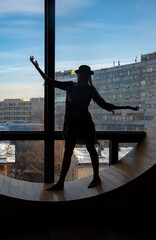 The silhouette of a girl in a dress and hat, standing at the window .