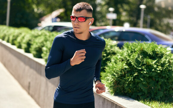 Horizontal image of an athlete male doing workout in the city street outdoors. Fit man in blue sports clothing and eyewear running with earphones outside on a sunny day.