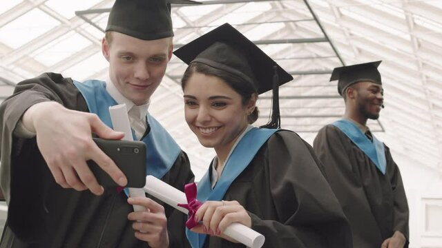 Tracking left of young mixed-race woman and her Caucasian male classmate wearing university graduate gowns and hats, holding diplomas and making selfie using phone camera