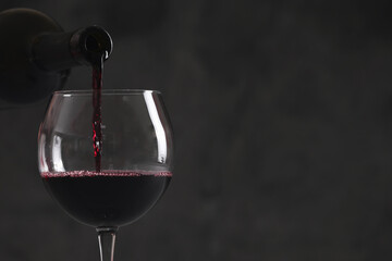Naklejka na ściany i meble Pouring red wine from bottle into glass on dark background, closeup. Space for text