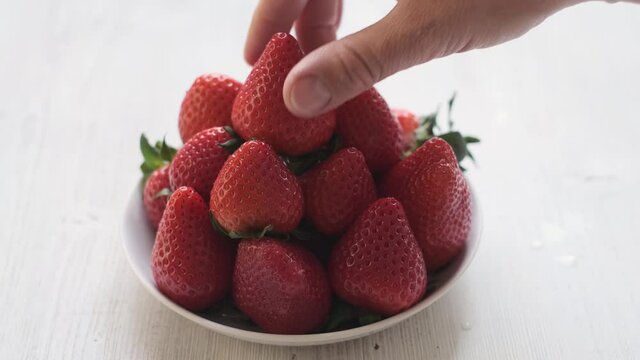 a hand placing a strawberry berry on a plate. Beautifully stacked red strawberries on a white wooden background.