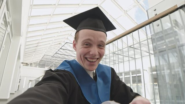 Medium shot of young Caucasian man wearing university graduate gown and hat making video call and showing his diploma to interlocutor during graduation day
