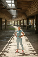 Fit girl standing on the side, hands on hips and looking at the camera while posing outside.