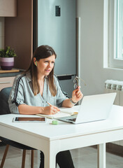 Businesswoman Working at Home on Laptop for Video Conference Call.
Serious business woman using laptop computer and wireless earphones to talking with coworkers on video call and writing down notes.