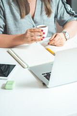 Unrecognizable Female Drinking Coffee and Working at Home on Laptop for Conference Call Meeting with Colleagues 
High angle view of woman sitting at desk with laptop, tablet, coffee and open notebook.