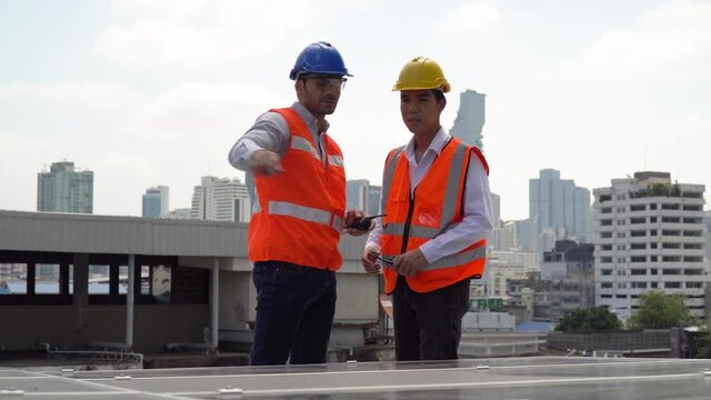 Solar power plant engineer manager with walkie talkie talking to asian worker control inspecting photovoltaic on rooftop in urban city. technician checking Solar Cell on roof outdoors on cityscape