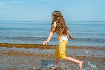 Fototapeta na wymiar A little girl in bright yellow pants runs in the water and frolics on the beach