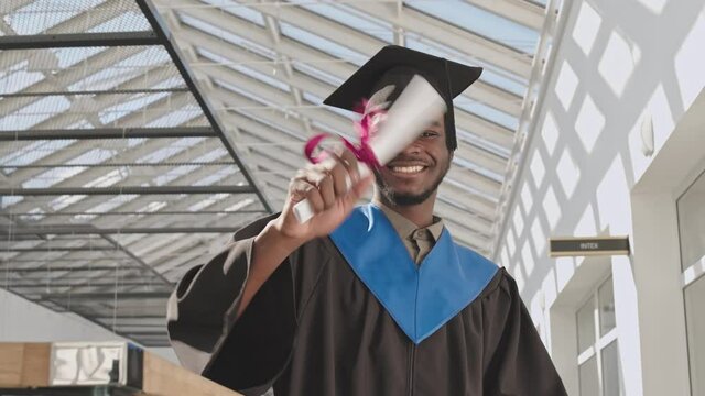 Panning of young African-American male student wearing university graduate gown and hat standing in university hall, looking at camera and waving his hand with diploma
