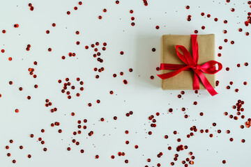 Gift in craft packaging with a red satin ribbon on a white background with chaotically scattered red shiny sequins. Gifts for Christmas, Valentine's Day, March 8. Horizontal photo. 
