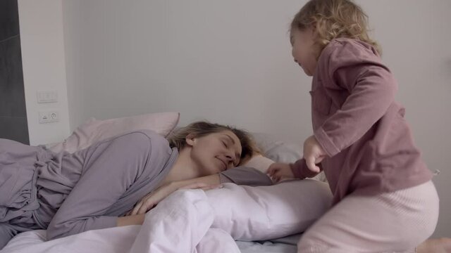 Little daughter wakes up her sleeping mom on the bed