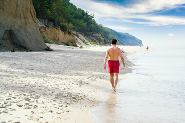 Young man in red shorts is walking on the water edge of Baltic sea. Beach next to the high cliff.