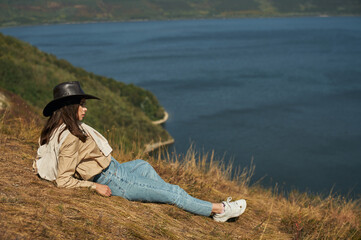 Charming young brunette in sensible clothes and cowboy hat lying on high hill and enjoying scenic landscape of Dniester canyon. Concept of recreation and nature.