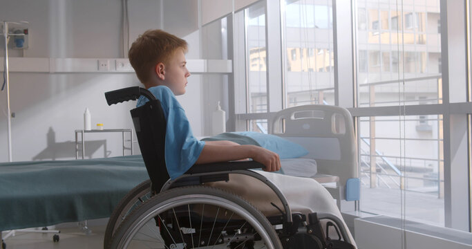 Side View Of Pensive Kid Sitting On Wheelchair Looking Out Of Window In Hospital Ward