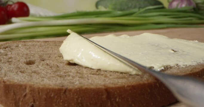 Butter Spread is Spreading on a Bread Slice with a Knife on a Cutting Board with Vegetables on the Background 
