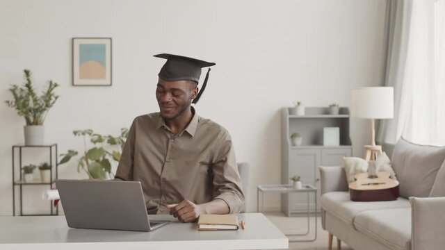 Medium shot of young African man wearing shirt and graduation cap sitting at desktop at home and having online graduation ceremony