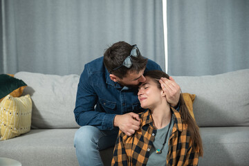 Fototapeta premium Young gentle man kissing his girlfriend in the forehead while she sitting on the floor and he on the sofa in their cozy apartment