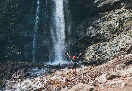  A woman dressed in a red hat and active trekking clothes standing near the mountain river waterfall rose arms up and enjoying the splashing Nature power. Traveling, trekking, and nature concept image