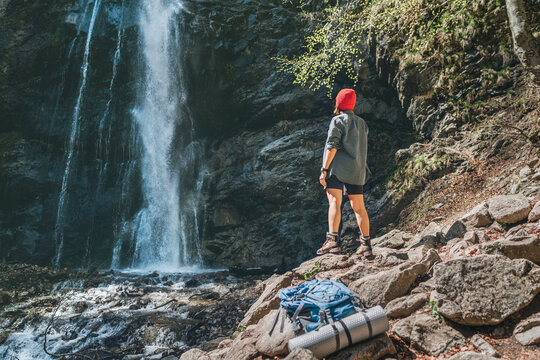  Woman with backpack dressed in red hat and active trekking clothes standing near the mountain river waterfall and enjoying the splashing Nature power. Traveling, trekking and nature concept image.