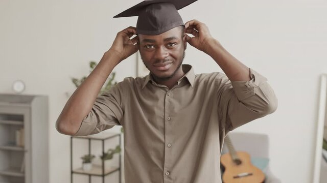 Medium shot of young African-American man standing in living room, putting on graduation hat while looking at camera, pointing at camera and saying joyful words