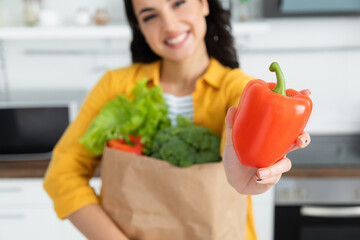 blurred and happy brunette woman holding paper bag with groceries and bell pepper