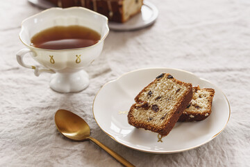 Slice of Pound Loaf Cake with raisins on white porcelain plate and cup of tea on greige linen tablecloth. Selective focus