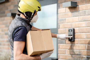Courier in green helmet wearing mask and gloves holds a parcel box and staying near front door of...