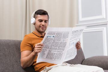 Naklejka premium positive man in wireless headphones reading newspaper on sofa at home