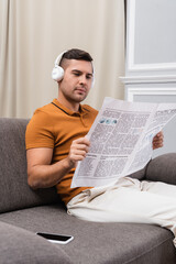 Fototapeta premium young man reading newspaper and listening music in headphones near smartphone with blank screen