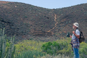 Fototapeta na wymiar Senior man hiking in arid landscape with mountain, cactus, holding backpack and binoculars
