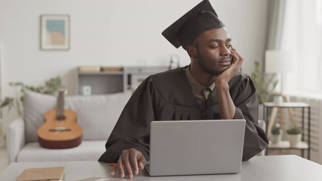 Medium shot of young handsome African man wearing graduation gown and cap sitting at desk in front of laptop at home and waiting for beginning of graduation ceremony