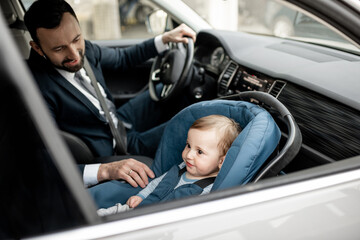 Father tend a child while driving at work with baby sitting in modern car seat. Child new born traveling safety on the road. Safe way to travel fastened seat belts in a vehicle with young kids. 