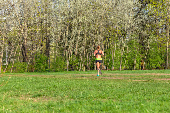 Young Sporty Tanned Girl Running In The Morning Green Park. A Woman In Short Sportswear Is Jogging While Listening To Music On Headphones. Morning Run. Girl Running From Behind