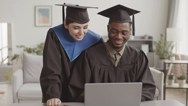 Medium shot of young mixed-race woman and African man both wearing graduation gowns and hats having online graduation ceremony at home using laptop