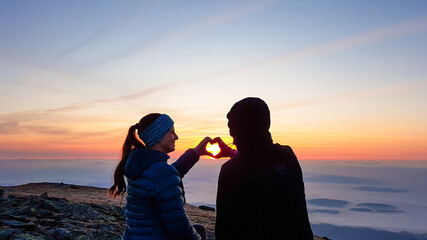 A couple sitting on top of Babia Gora, Poland, and enjoying the panoramic view on sun rising above the horizon. They form a heart with their hands. Thick clouds below. The sky is pink and orange. Love