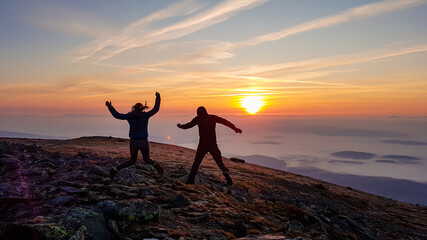 A couple holding hands and jumping on top of Babia Gora, Poland, with the panoramic view on sun rising above the horizon. Thick clouds below. The sky is pink and orange. Love and passion