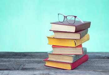 Glasses and a stack of six multi-colored books lie on a wooden table on a turquoise background