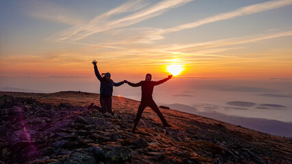 A couple holding hands and jumping on top of Babia Gora, Poland, with the panoramic view on sun...