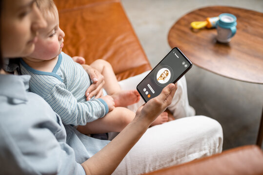 Young Mother With Newborn Kid Making Video Call To A Doctor Using Phone At Home. Concept Of Telemedicine And Support Parents Counseling Online. 