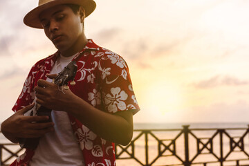 young Latin American with a Hawaiian shirt and a hat holding a stringed instrument.
