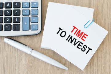 TIME TO INVEST is written on a white note card on a wooden background near the calculator and pen. Investment concept