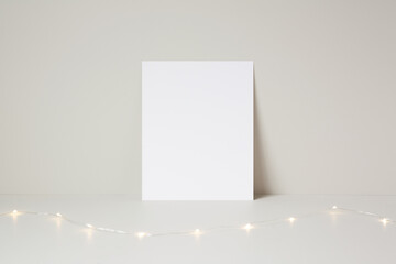 Festive vertical blank paper mockup template resting against a light brown background wall, with fairy lights prop.