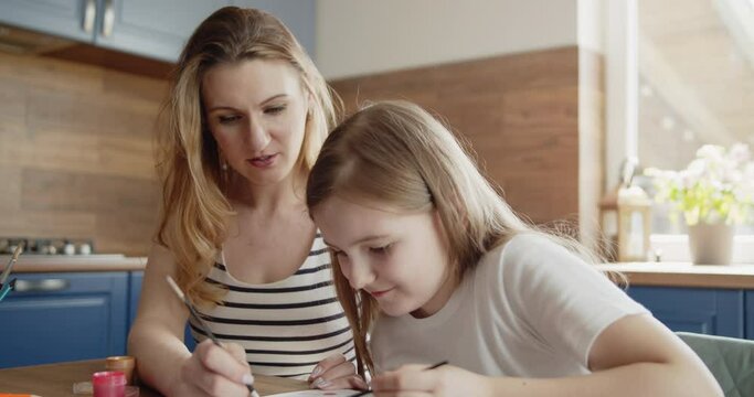Mother and daughter draw a picture with paint on paper sitting together by the table in the kitchen. Young mom helps her kid to paint a picture. Lifestyle, hobbies, leisure, arts and education concept