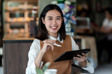 Happy young Asian woman holding a tablet with a pen at café. Looking at the camera.
