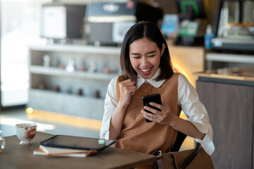 Front view young Asian woman sitting at the coffee shop watching the smartphone rejoicing success.