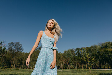 Young woman with long blonde hair, wearing a blue dress, posing on blue sky background, outdoors.