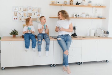 Mom with her two children sitting on the kitchen table. Mother with daughter and toddler son having breakfast at home. Happy lifestyle family moments.