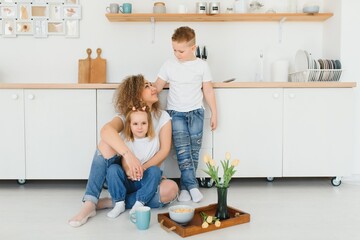 Happy mother hugging daughter and son sitting on wooden floor in modern kitchen at home. Mom hugging children. Happy family relationship concept