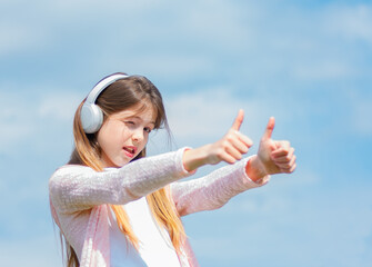 cute playful girl wearing headphones and showing thumbs up on hands on sky background